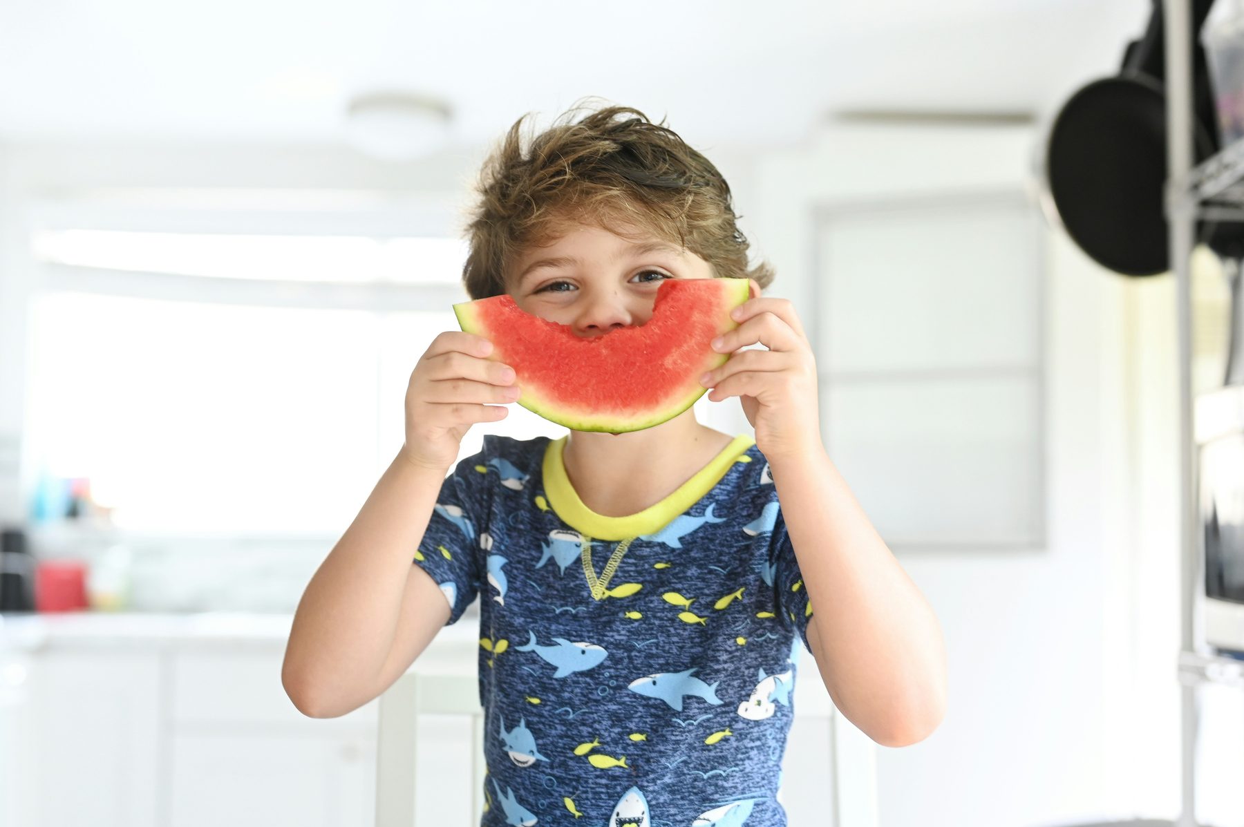 A child holding a slice of watermelon over their face in a bright kitchen
