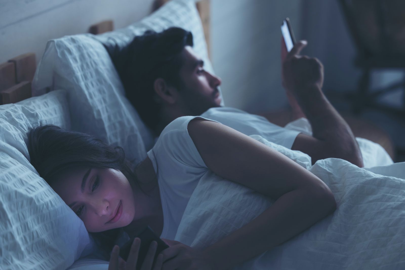 Two adults lying in bed at night, both scrolling their phones