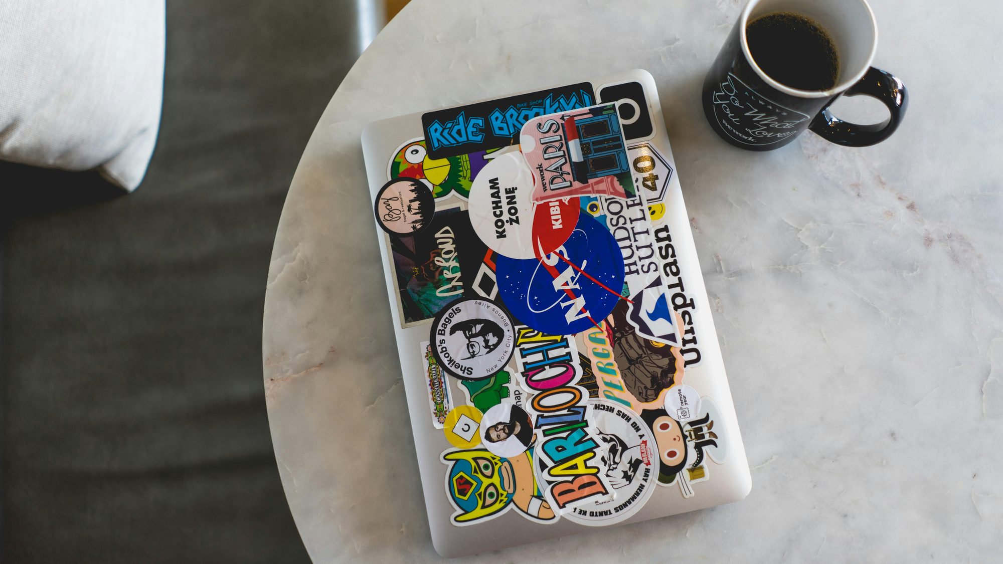 A sticker-covered laptop closed on a table beside a cold mug of coffee