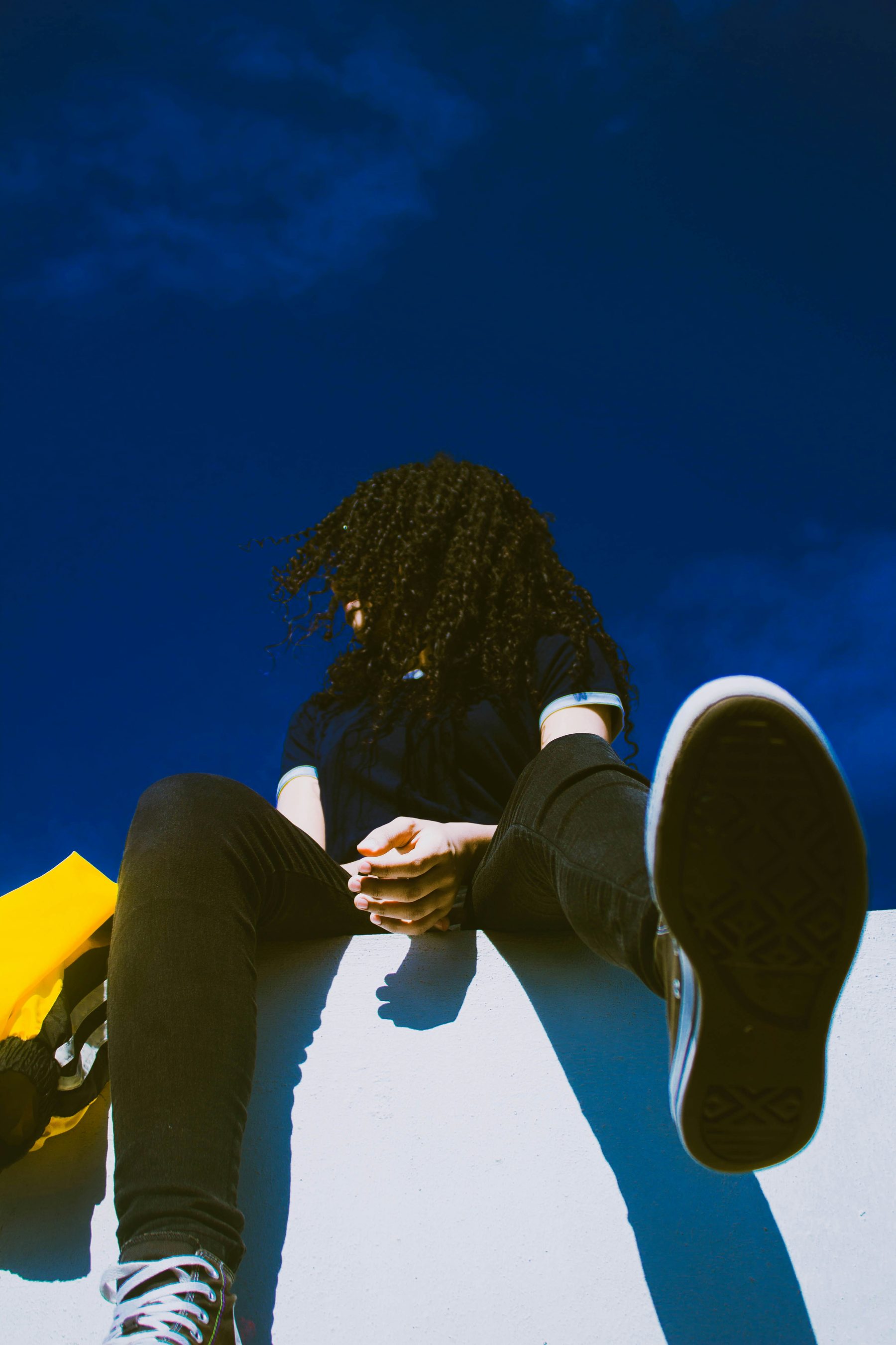 A teenager sitting on a ledge against a blue sky, looking at their phone
